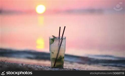 Close up of glass of mojito alcoholic cocktail standing in the sand on a tropical beach at sunset in the evening against a solar disk moving down