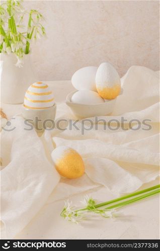 Close up of golden and white Easter eggs with white flower on bright white wooden table background.