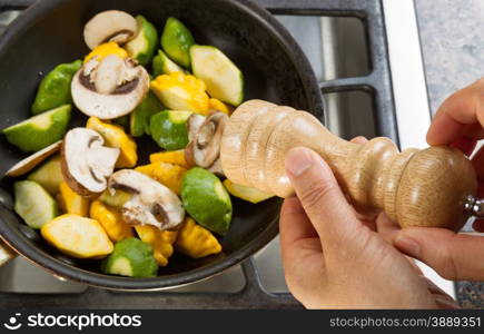 Close up of hand, focus on pepper shaker, holding a pepper shaker with frying pan filled with vegetables underneath.