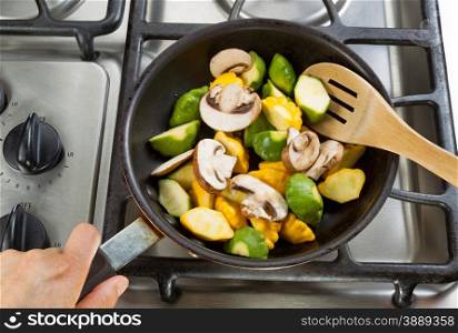 Close up of hand holding frying pan, focus on food, while stirring vegetables with wooden spoon.