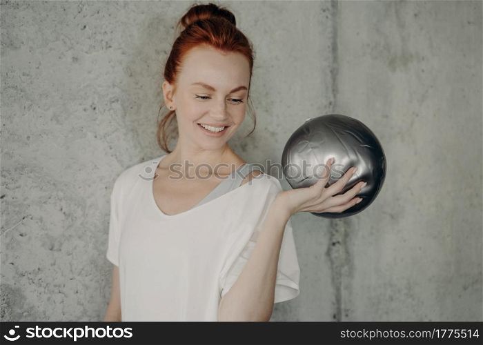 Close up of happy ginger woman against blurred concrete wall background in fitness studio looking on silver barre fitball in her hand and smiling, female enjoying pilates class in gym. Smiling redhead woman with silver fitball in her hand during pilates workout