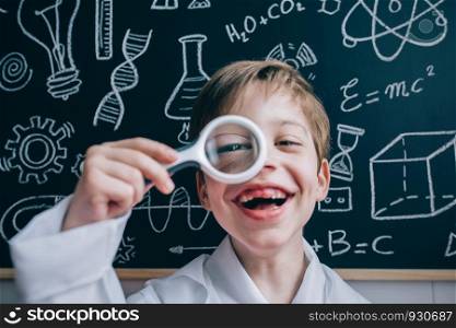 Close up of happy little scientist laughing and looking at camera while looking through magnifier against of drawn blackboard. Happy kid looking at camera through magnifying glass
