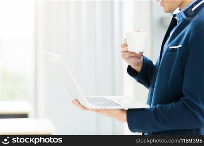Close-up of Happy mood asian young businessman working with laptop computer and holding a coffee cup at the window In the office room background.