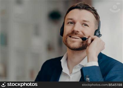 Close up of happy office worker handsome male call center operator wearing headset and working in office, talking with client online with positive face expression, selective focus. Job and occupation. Happy young man consulting client by call