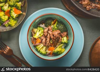 Close up of healthy balanced nutrition meal in bowl with beef meat, rice , steamed vegetables: broccoli and carrots served with plate and cutlery, top view