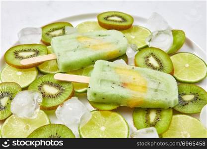 Close-up of healthy home-made fruit ice cream on a stick with a slice of peach in a plate with pieces of lime, kiwi and cold ice cubes. Organic dietary dessert. Close-up of homemade lime kiwi icy popsicle with a piece of peach in a plate with ice cubes and pieces of fruit