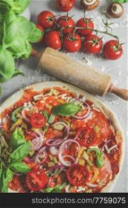 Close-up of homemade pizza with tomatoes, salami, onion and basil at kitchen table with flour, wooden rolling pin, basil and ingredients. Cooking traditional Italian food. Top view.
