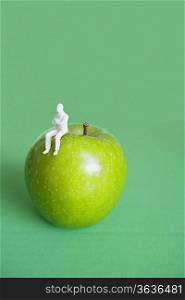 Close-up of human figurine sitting on green apple over colored background