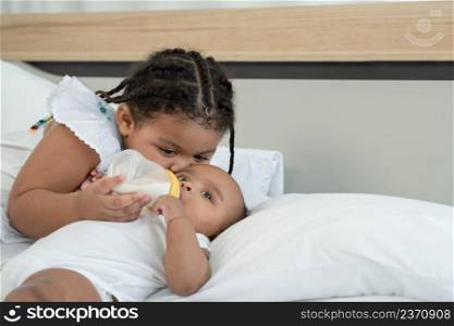 Close up of little adorable African older sister feeding newborn baby milk bottle and kissing with love and care while infant lying on bed at home. sisterhood, child care by siblings concept