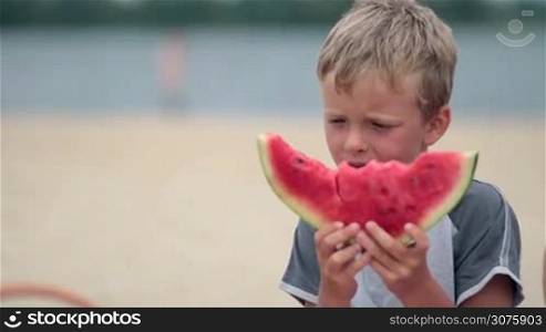 Close up of little boy eating delicious watermelon and licking his lips on the beach in summertime. Blurred river bank on background