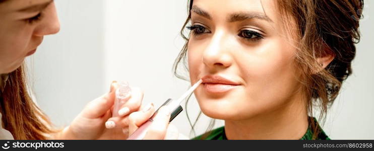 Close up of makeup artist applying lipstick foundation with a make-up brush on lips of the beautiful young caucasian woman in a beauty salon. Makeup artist applying lipstick foundation