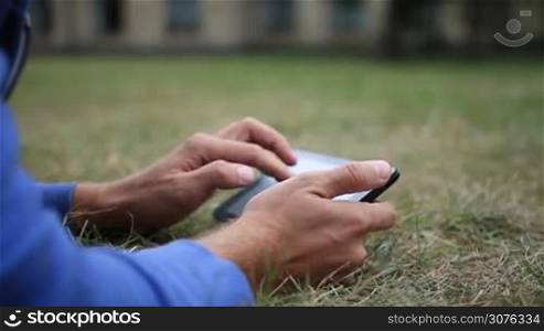 Close up of male hands with digital tablet touching the screen. Young student lying on the grass in the park ouside the university and learning online using a tablet.