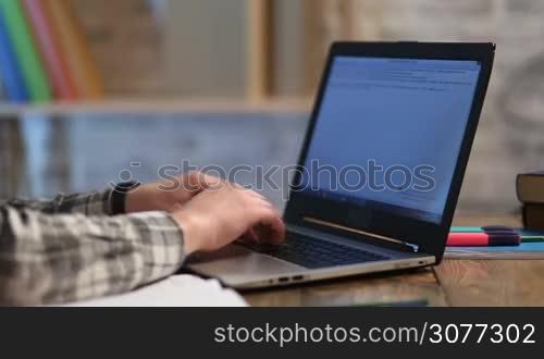 Close up of male student hands typing text on modern laptop on wooden table while studying at home. Young teenager working on computer in library and preparing for exams. Hipster using digital gadget.