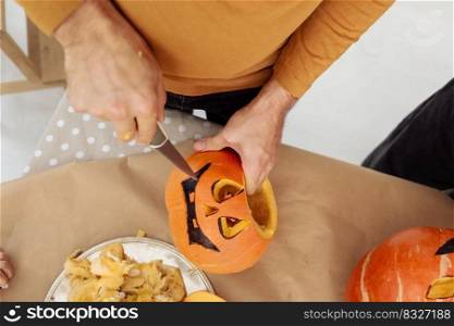 Close up of man hands carving Jack O Lattern from ripe orange pumpkin with knife on her wooden kitchen table. male preparing all hallows eve Halloween party decorations. Background, copy space. Close up of man hands carving Jack O Lattern from ripe orange pumpkin with knife on her wooden kitchen table. male preparing all hallows eve Halloween party decorations. Background, copy space.