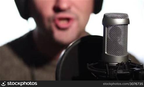 Close up of man singing into a condenser microphone in studio against white background. Male vocalist in headphones singing into microphone in recording studio.