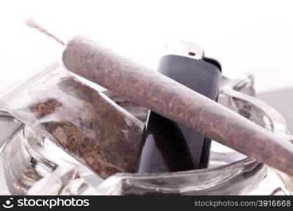 Close up of marijuana joint made with translucent rolling papers, plastic baggy of dried marijuana, black lighter and pipe on white background