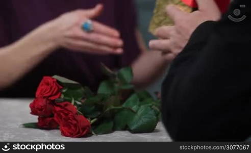 Close up of mature man&acute;s hands giving golden gift box with red ribbon to his beloved woman during celebration of Valentine&acute;s day. Elderly husband giving present to wife on anniversary at the restaurant over table decorated with bouquet of red roses.