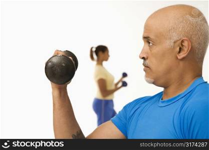 Close up of mid adult multiethnic man exercising using dumbbells with mid adult multiethnic woman in background.