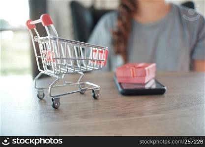 Close up of miniature shopping cart /trolley toy figure. smart phone and red gift box on table for shopping and e-commerce concept