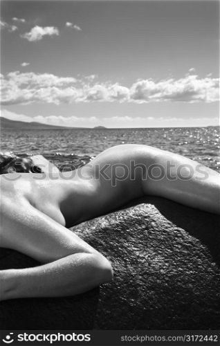 Close up of nude Caucasian mid adult woman draped over rock with at Maui coast in background.