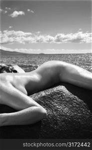 Close up of nude Caucasian mid adult woman draped over rock with at Maui coast in background.