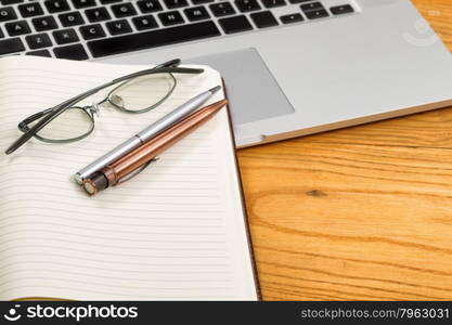 Close up of pens, selective focus on pens, with open blank notebook, reading glasses, and computer in background on desktop.