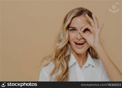 Close up of positive young woman with long wavy fair hair looking at camera through fingers folded into ok gesture, opens mouth with excitement while posing isolated over beige background. Positive young woman looking at camera through fingers folded into ok gesture