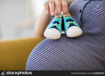 Close Up Of Pregnant Woman Sitting On Sofa At Home Holding Baby Shoes On Stomach