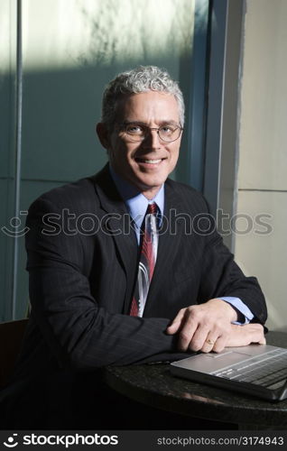 Close up of prime adult Caucasian man in suit sitting at table with laptop looking at viewer and smiling.