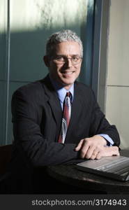 Close up of prime adult Caucasian man in suit sitting at table with laptop looking at viewer and smiling.