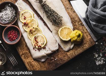 Close up of raw cod fillet with lemon slices and herbs on rustic wooden cutting board, top view. Fish cooking preparation. Healthy diet food