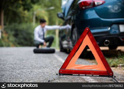 Close up of Red triangle sign behind the broken car. Side view of Businessman sitting on road sign wheel changing. Warning triangle in front of broken-down car on the road.