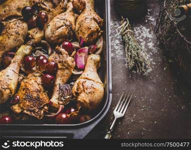 Close up of roasted chicken drumsticks with red onion and grapes on dark background.