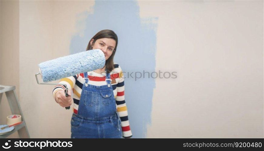 Close-up of roller with blue paint in the hand of a standing woman dressed in overalls and striped blouse, seen from the front, against background of half-painted white wall with strokes of blue paint. roller with blue paint in the hand of a standing woman dressed in overalls and striped blouse, seen from the front, against background of half-painted white wall with strokes of blue paint