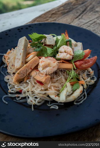 Close-up of Seafood spicy glass noodle salad or Spicy delicious Mung bean Noodle Salad with fresh seafood (Yum woon sen) on wooden table. Spicy vermicelli salad, Vegetables and herbs, Sour & spicy, Thai cuisine, Selective focus.