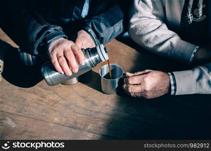 Close up of senior couple hands pouring coffee in a metal cup from thermos over a wooden table. Senior couple hands pouring coffee from thermos