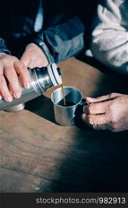 Close up of senior couple hands pouring coffee in a metal cup from thermos over a wooden table. Senior couple hands pouring coffee from thermos