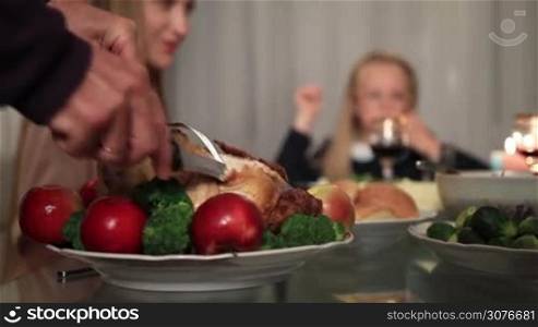 Close up of senior male hand carving slices of roasted turkey with apples and broccoli on platter over blurred family sitting at festive table decorated with candles background during Thanksgiving celebration.