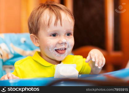 Close-up of shot of a happy cute boy sitting at feeding table, his face and hands dirty with cream yoghurt