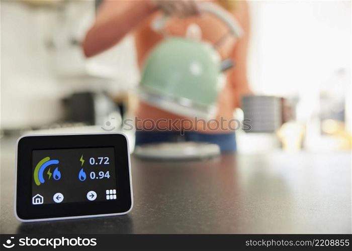 Close Up Of Smart Energy Meter In Kitchen Measuring Electricity And Gas Use With Woman Boiling Kettle