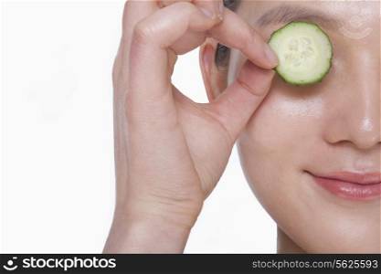 Close up of smiling young woman holding up a cucumber slice over her eye, studio shot