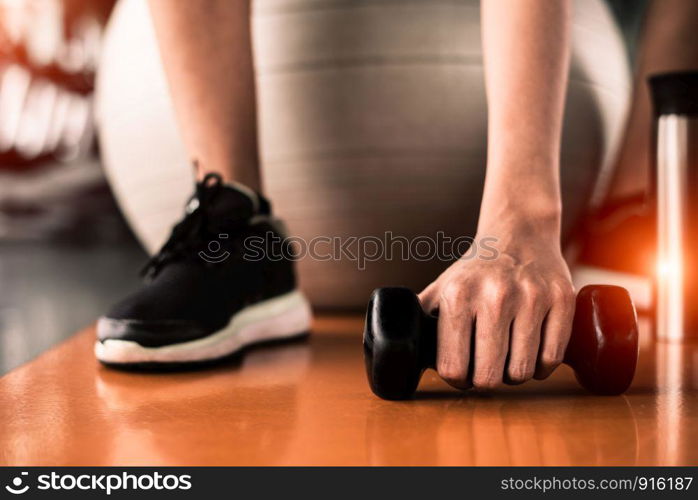 Close up of sport woman sitting on yoga ball and grab dumbbell on floor by hand in fitness sport club center background. Sport and recreation activity concept. Weight training and bodybuilding concept