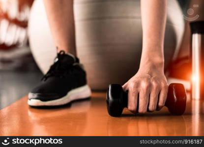 Close up of sport woman sitting on yoga ball and grab dumbbell on floor by hand in fitness sport club center background. Sport and recreation activity concept. Weight training and bodybuilding concept