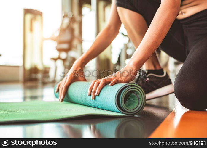 Close up of sporty woman folding yoga mattress in sport fitness gym training center background. Exercise mat rolling keeping after yoga class. Workout and sport training concept. Hands on carpet