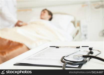 Close-up of stethoscope and medical chart with a patient lying in a hospital bed in the background