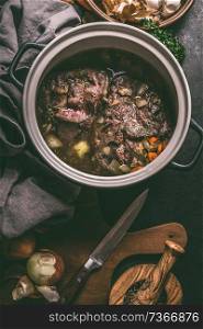 Close up of stewed beef meat in cooking pot with herbs, spices and kitchen utensils, top view. Country&rsquo;s meat dishes. Slow cooking. Rustic food