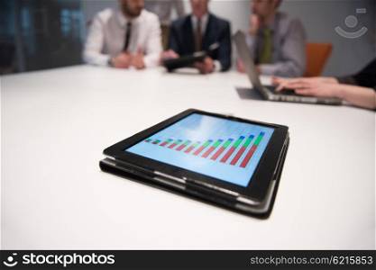 Close up of tablet touchpad computer with focus on business analytics and progress chart document. Business people group on meeting in background