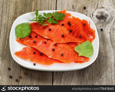 Close up of thinly sliced cold smoked red salmon, selective focus on front part of salmon, along with herbs and seasoning in white plate. Rustic wood underneath.