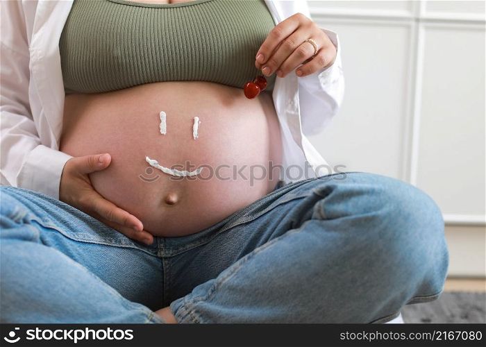Close-up of torso of young pregnant model applying moisturizer on her belly to avoid stretch marks. Future mom with funny smile from moisturizing cream on her tummy. Pregnancy skincare concept.. Close-up of torso of young pregnant model applying moisturizer on her belly to avoid stretch marks. Future mom with funny smile from moisturizing cream on her tummy. Pregnancy skincare concept