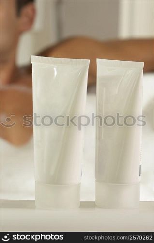 Close-up of two bottles of moisturizer with a young man in a bathtub behind it